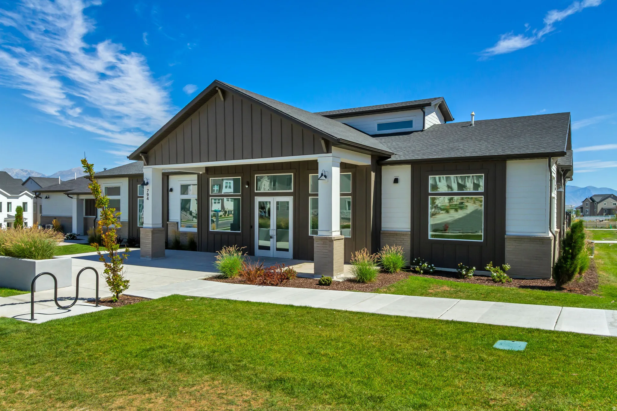 The community clubhouse boasts a modern exterior featuring vertical dark siding and white horizontal siding, a covered entry with stone-clad columns, large windows, and a manicured lawn with a concrete walkway