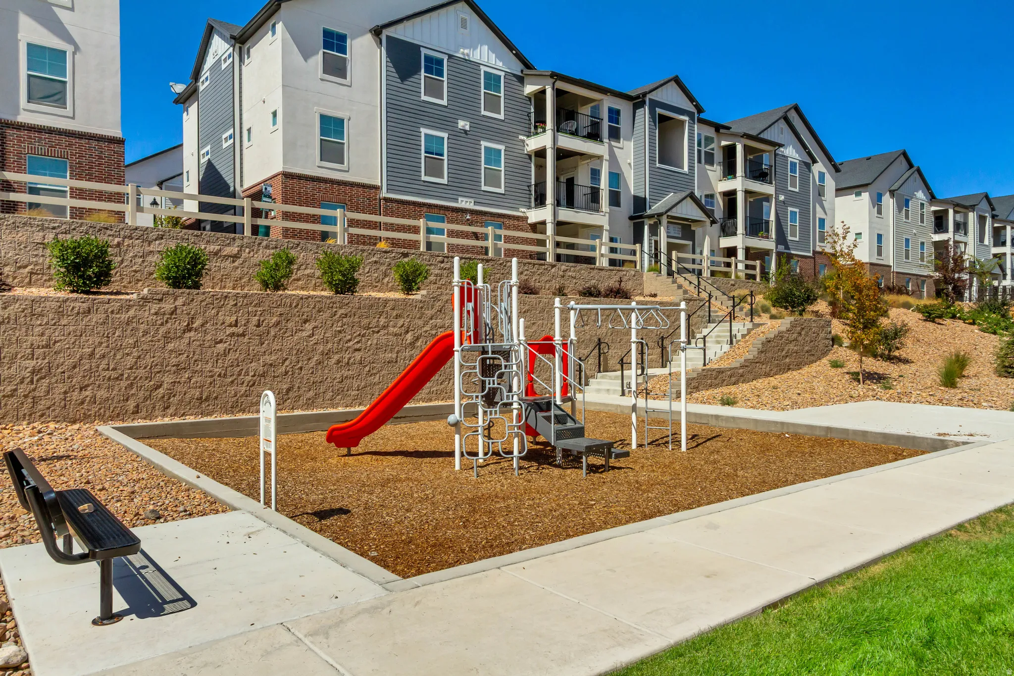 Outdoor playground with a red slide, a bench, a concrete walkway, and landscaped slopes