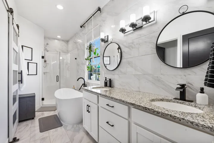 Dual vanity with granite countertop, freestanding soaking tub, glass-enclosed shower, barn door entry, and tiled flooring