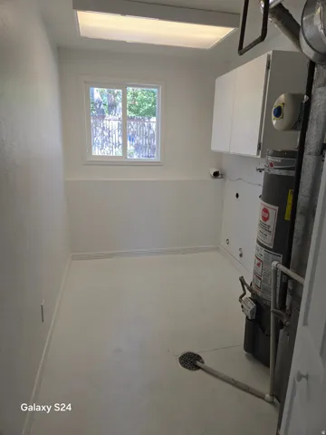 White cabinetry, fluorescent overhead lighting, window with exterior view, water heater, and floor drain