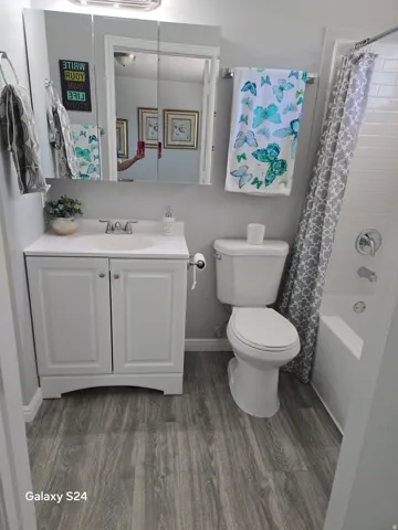 Bathroom featuring a white vanity with a built-in sink, wood-finish flooring, a wall-mounted medicine cabinet with integrated lighting, a toilet, and a white bathtub with subway tile surround