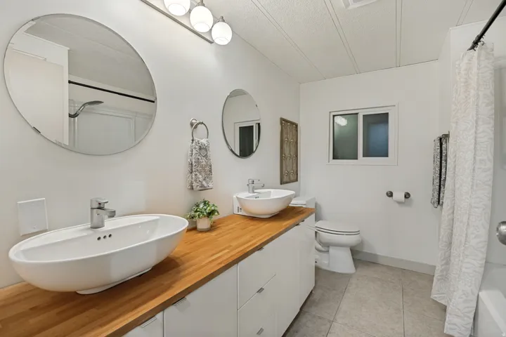 Hallway Bathroom. Dual vessel sinks set atop a wood-finish countertop, complemented by white cabinetry and a mounted light fixture