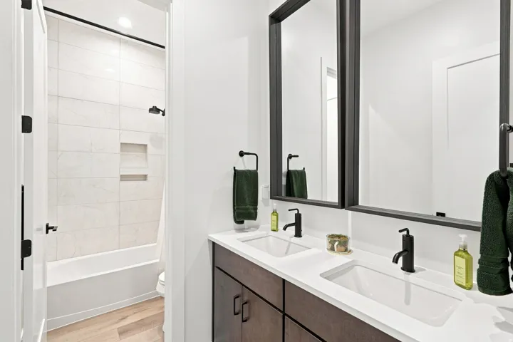 Dual vanity with integrated sinks and matte black fixtures, framed mirrors, wood-finish flooring, and a shower-tub combination with tiled surround and built-in niche