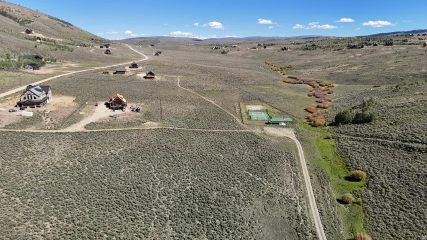 Expansive open land featuring a tennis court, a natural watercourse with green vegetation, a winding unpaved road, and distant mountain slopes