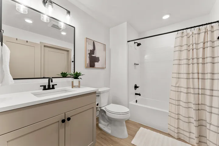 Light wood-finish flooring, a white vanity with a rectangular undermount sink, black-finish hardware, a white subway tile shower surround, and a ceiling-mounted light fixture