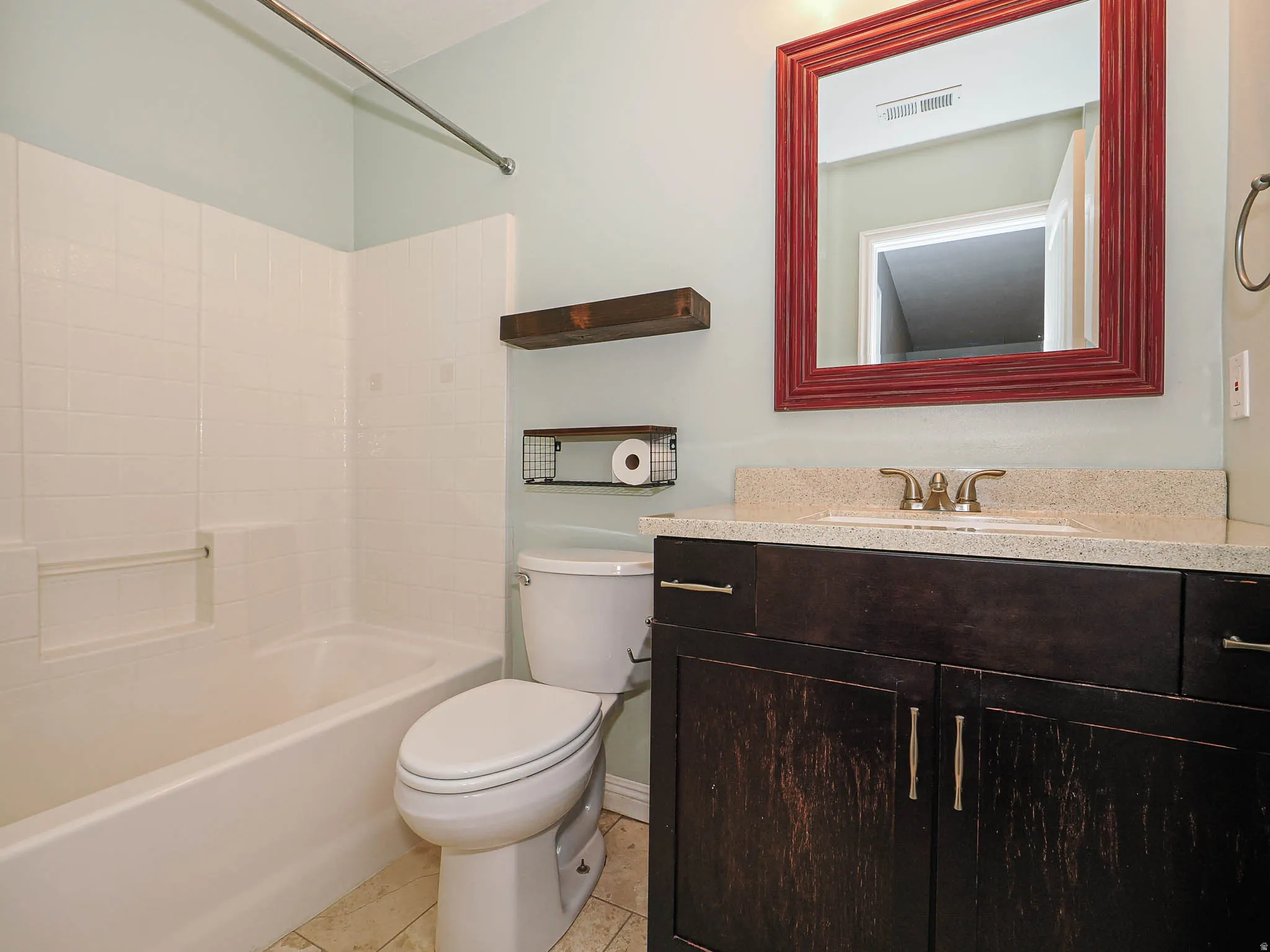 White bathtub with integrated shower, dark wood-finish vanity with an undermount sink, solid surface countertop, and a red-framed mirror