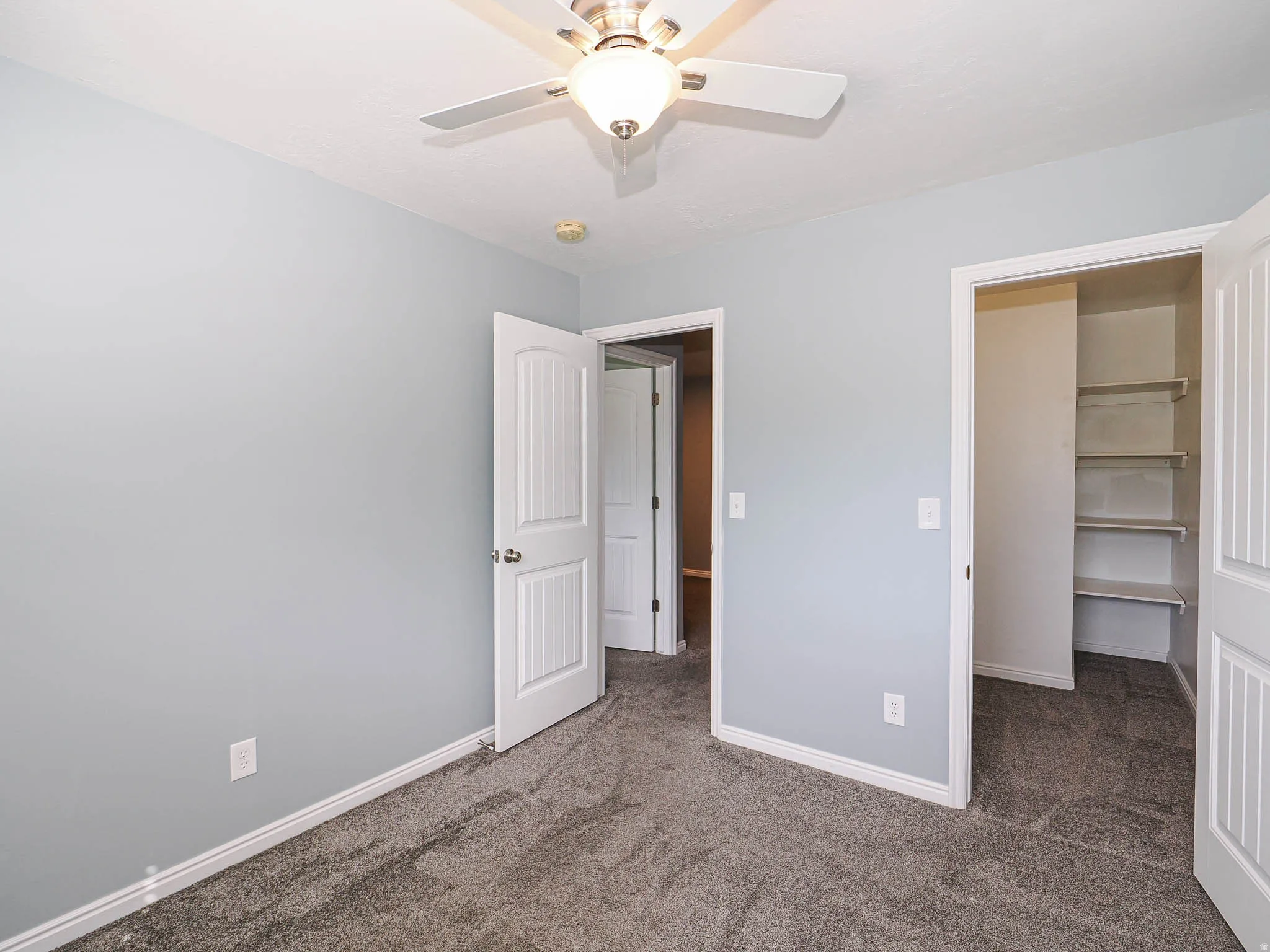 Light gray wall paint, textured gray carpeting, white trim, ceiling fan with integrated lighting, and a built-in closet storage system