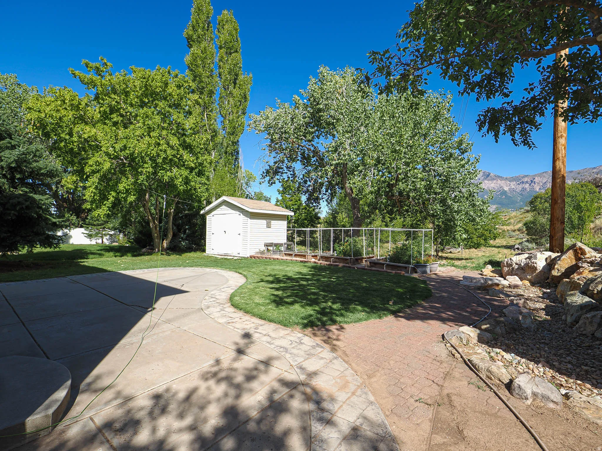 Expansive yard with concrete patio, brick paver pathway, storage shed, raised garden beds, and mountain scenery