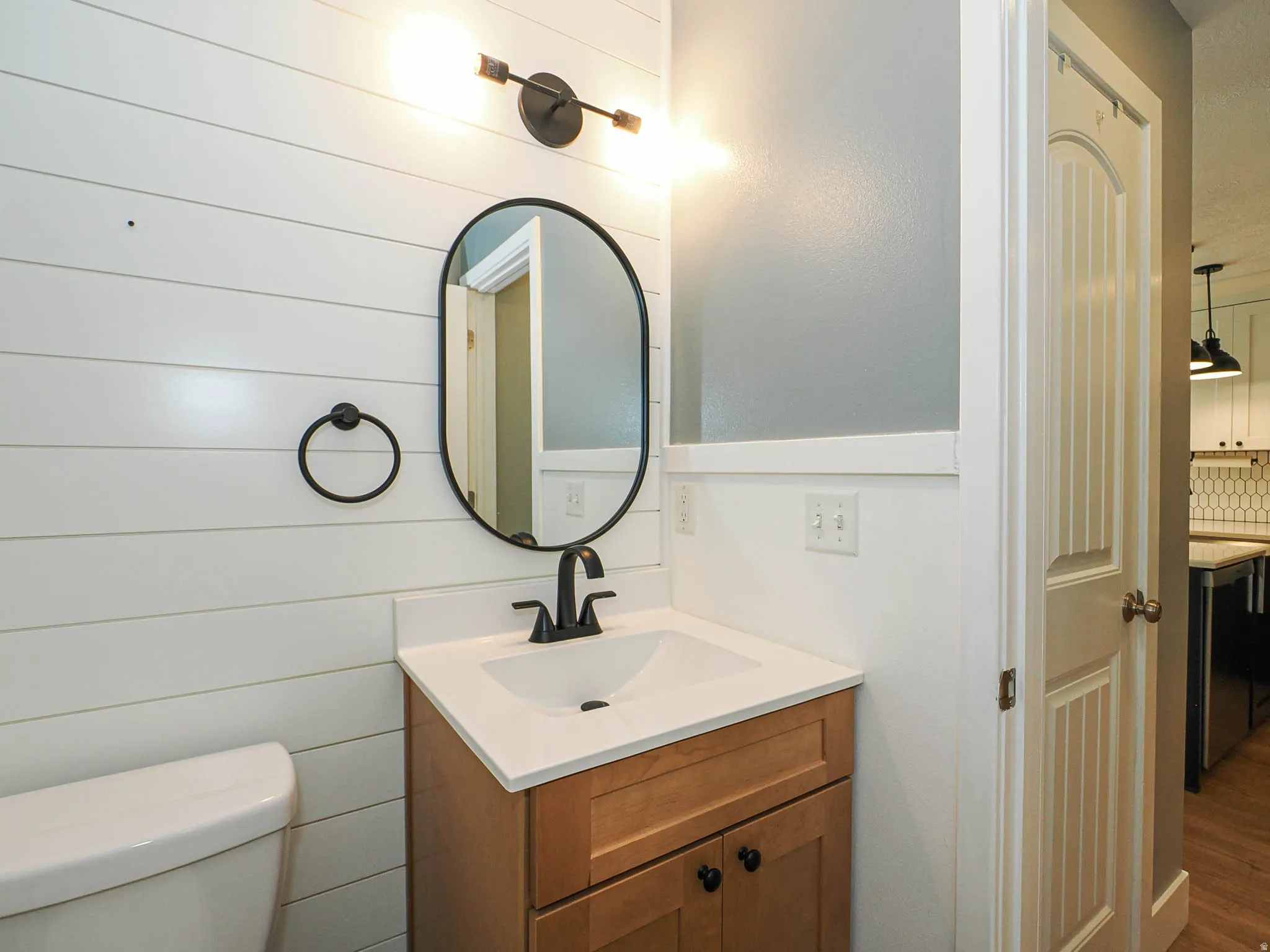 White shiplap wall accentuating the vanity with a black matte faucet, oval mirror, and a modern light fixture