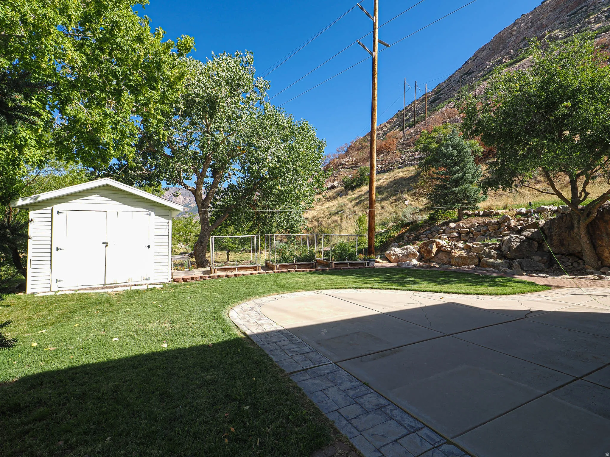 Landscaped yard featuring a white storage shed, mature trees, and a concrete patio with a decorative paver border