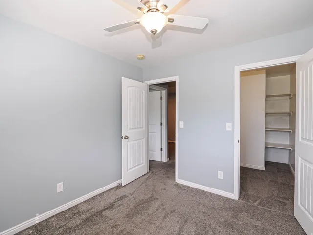 Light gray wall paint, textured gray carpeting, white trim, ceiling fan with integrated lighting, and a built-in closet storage system