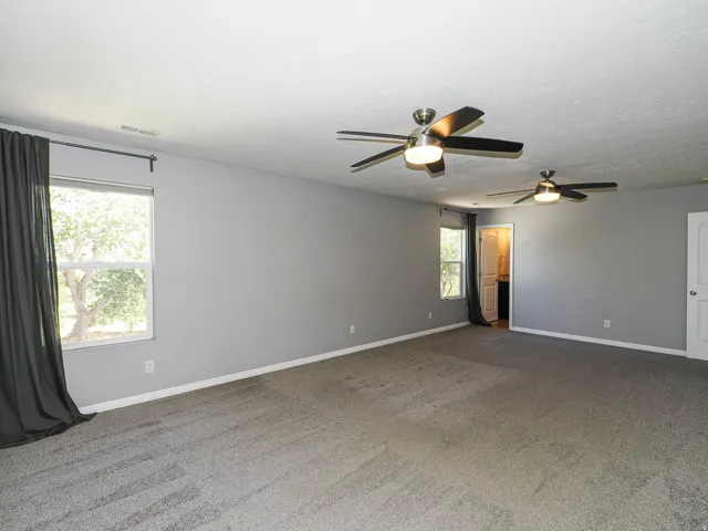 Spacious room featuring neutral carpeting, two windows, two ceiling fans with integrated lighting, and a white paneled door