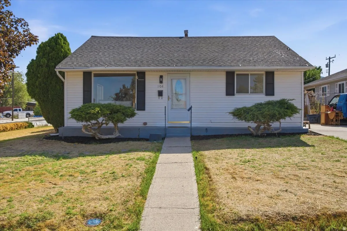 White siding exterior with a dark shingle roof, featuring a bay window, a concrete walkway, and established front landscaping
