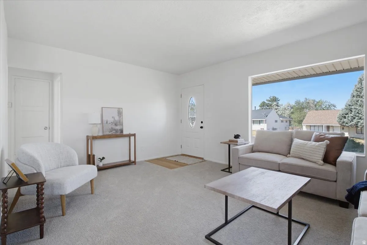 Bright living area with a large picture window, light-toned carpeting, and a white entry door with an oval glass insert