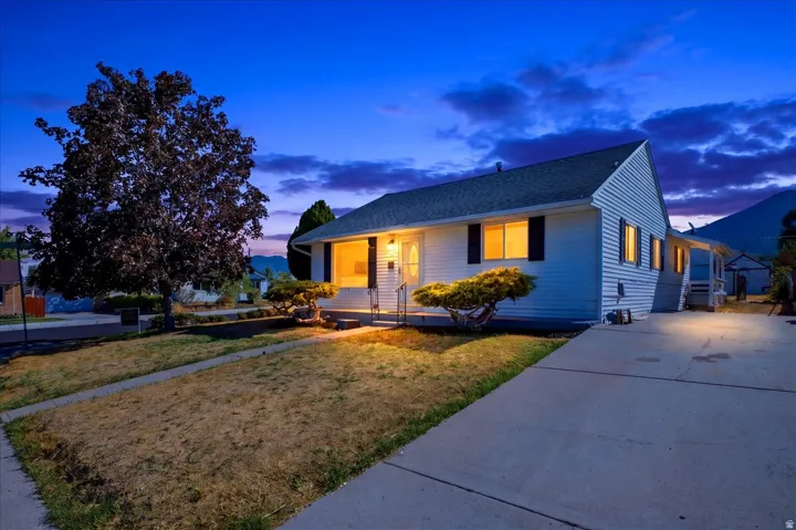 White siding exterior with black shutters, mature tree, established shrubbery, concrete driveway, and covered entry