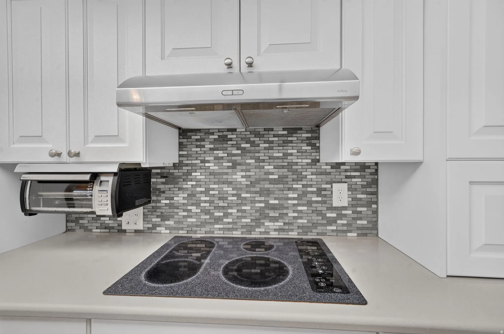 White paneled cabinetry with brushed nickel hardware, mosaic tile backsplash, built-in cooktop, and a stainless steel range hood