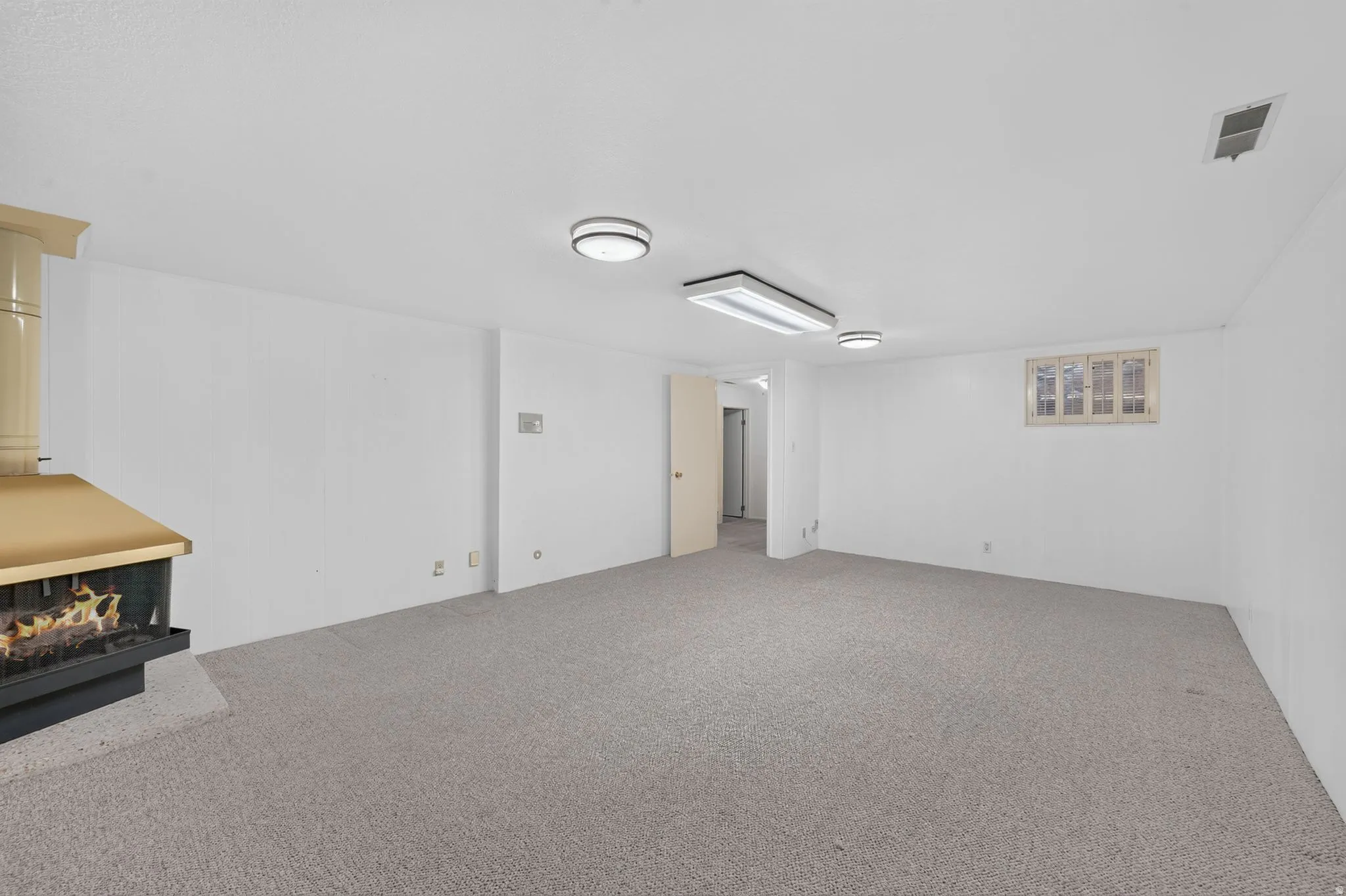 Spacious carpeted room featuring a freestanding fireplace with a metal hood, overhead flush-mount lighting, and recessed window