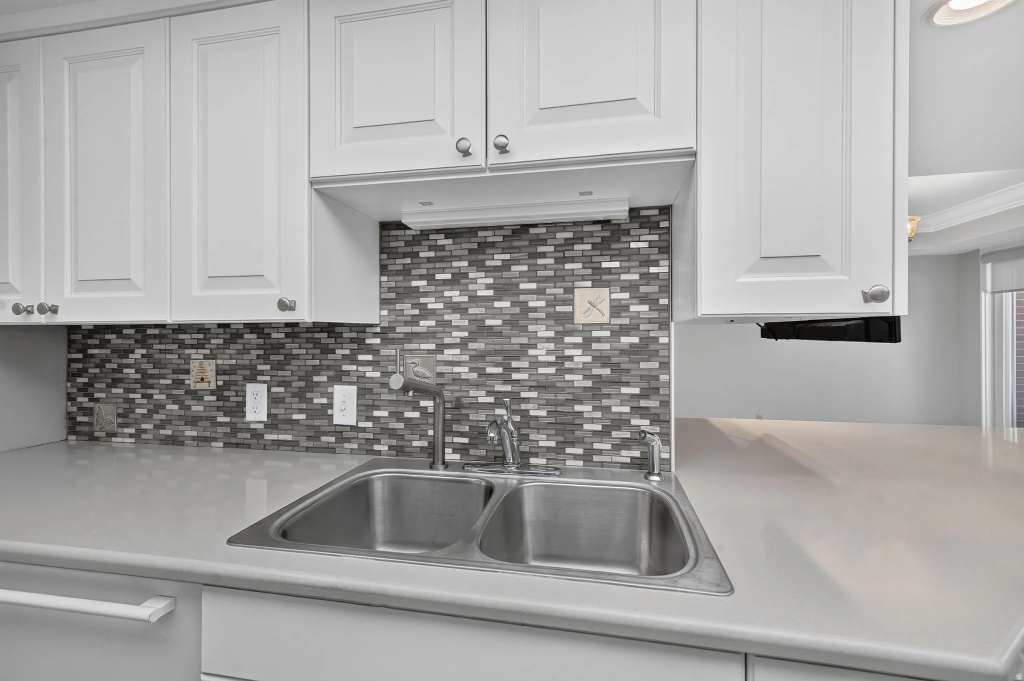 White cabinetry with silver-tone hardware, mosaic tile backsplash, double basin stainless steel sink, and light-toned countertop