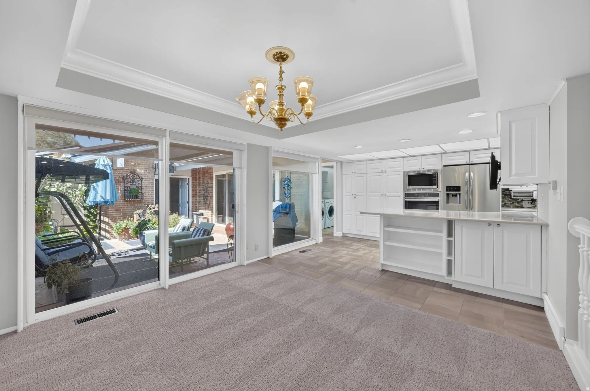 Tray ceiling with crown molding, brass chandelier, sliding glass doors, white cabinetry, and stainless steel appliances