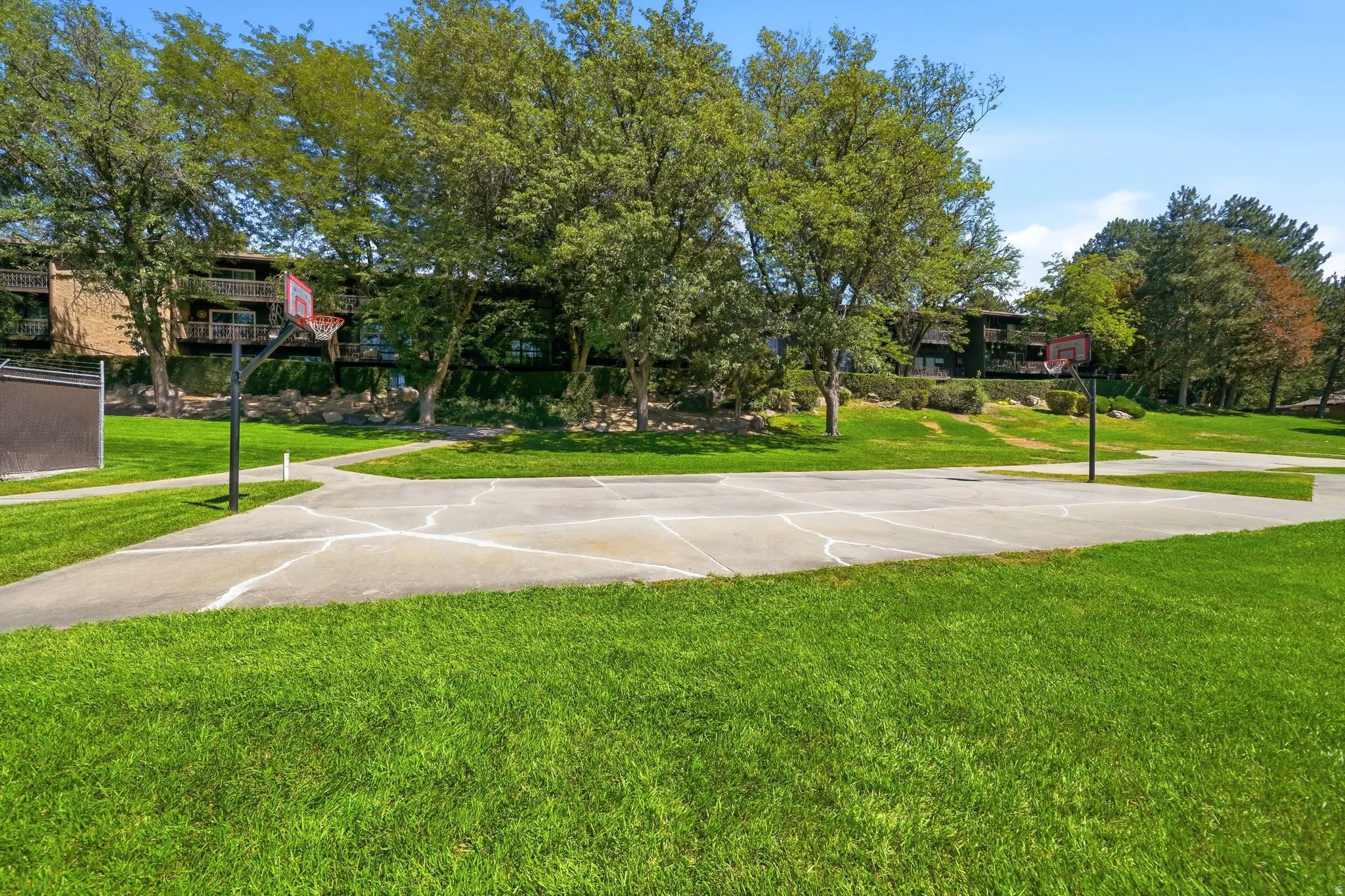 Basketball court with two hoops, surrounded by expansive green lawn, mature trees, and a paved pathway