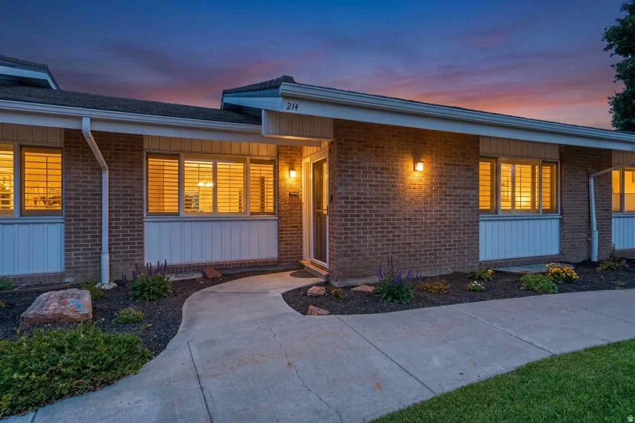 Brick exterior with white trim, a curved concrete pathway, established landscape plantings, and exterior wall-mounted lighting