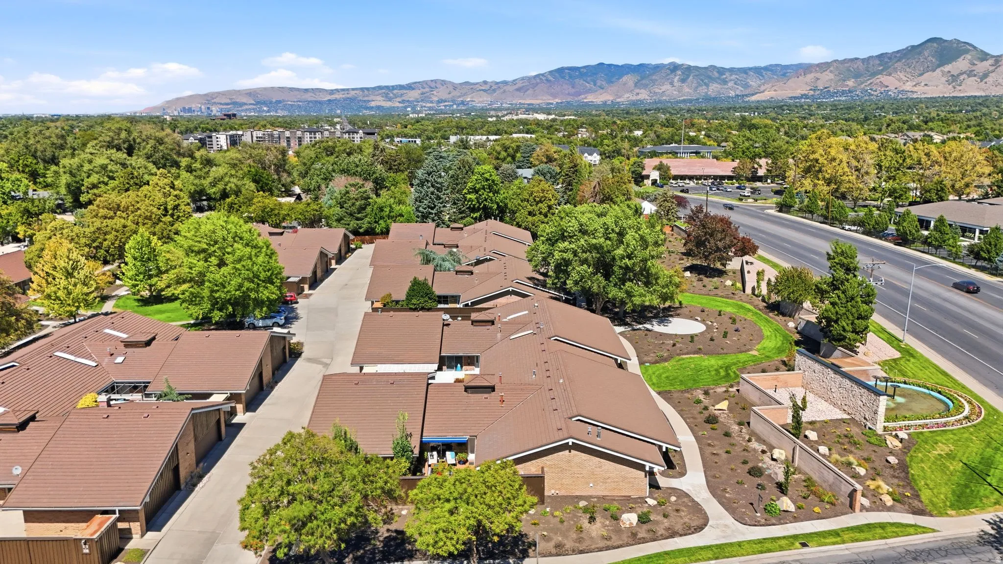 Expansive mountain backdrop, community landscaping, mature trees, brown shingle roofs, and a multi-lane road