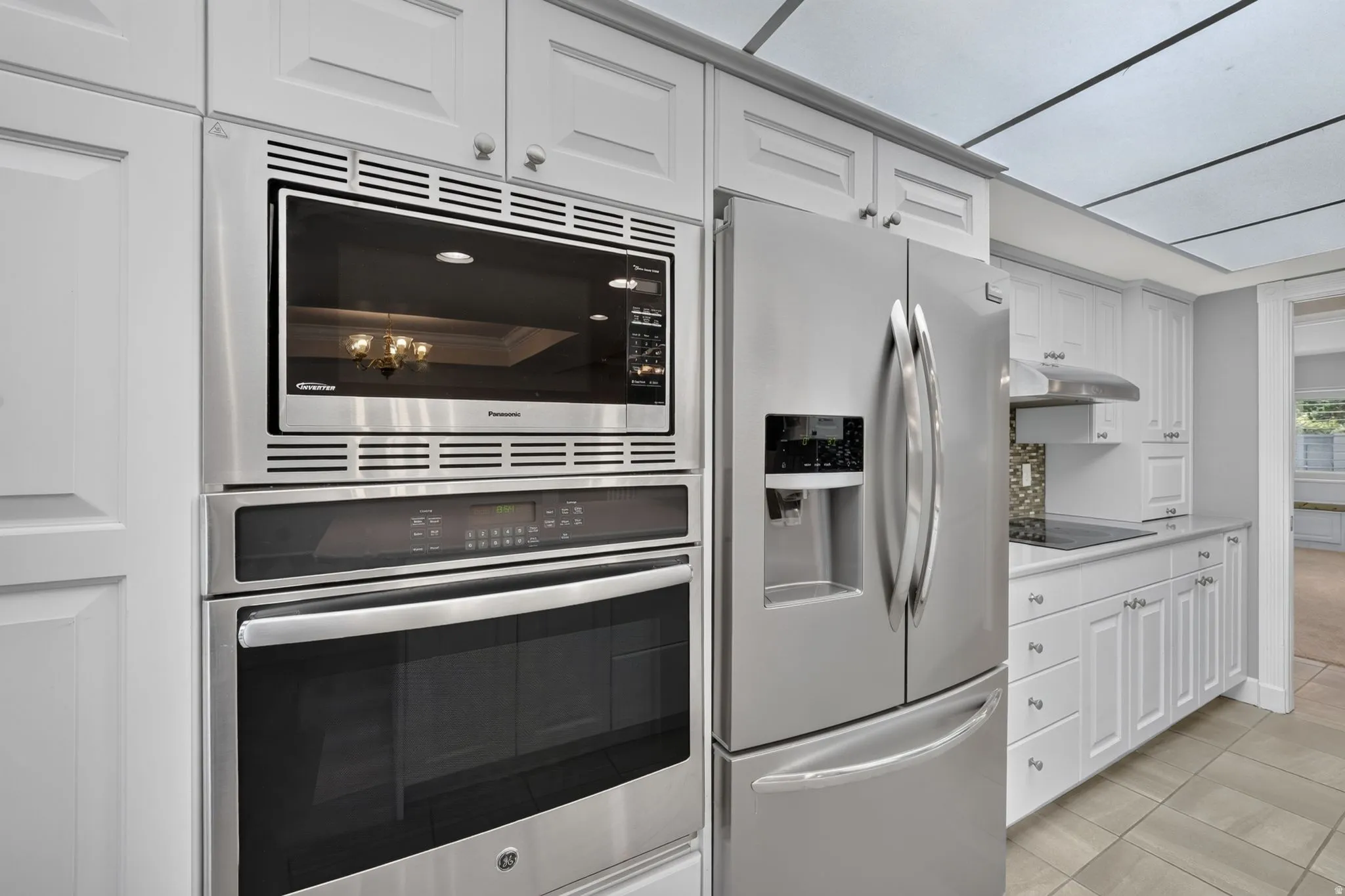 White shaker-style cabinetry with chrome hardware, stainless steel built-in microwave and oven, stainless steel French door refrigerator, light-toned tile flooring, and a tile backsplash