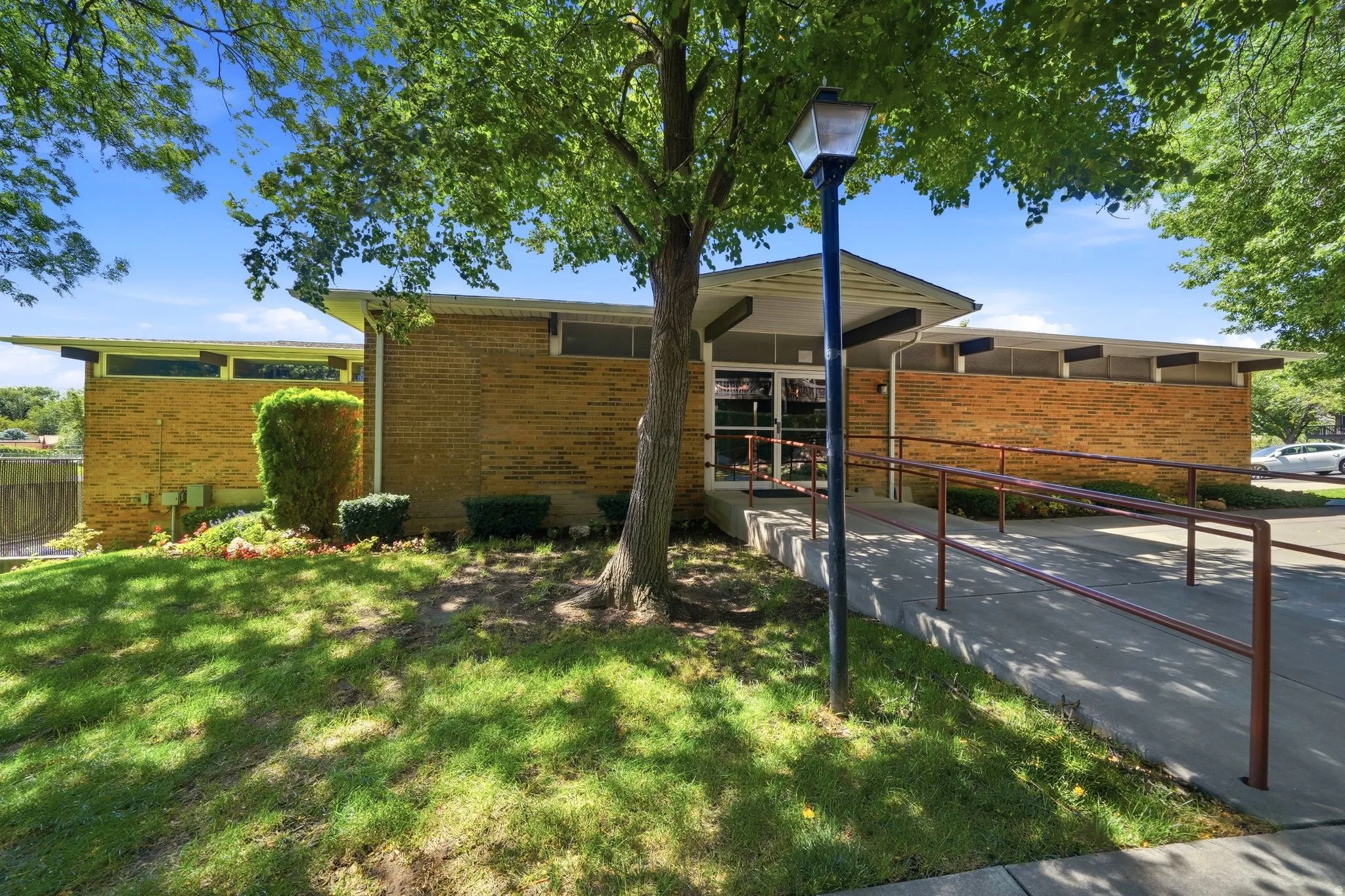 Brick exterior with high windows and a covered entry, featuring a concrete ramp with metal handrails and established landscaping