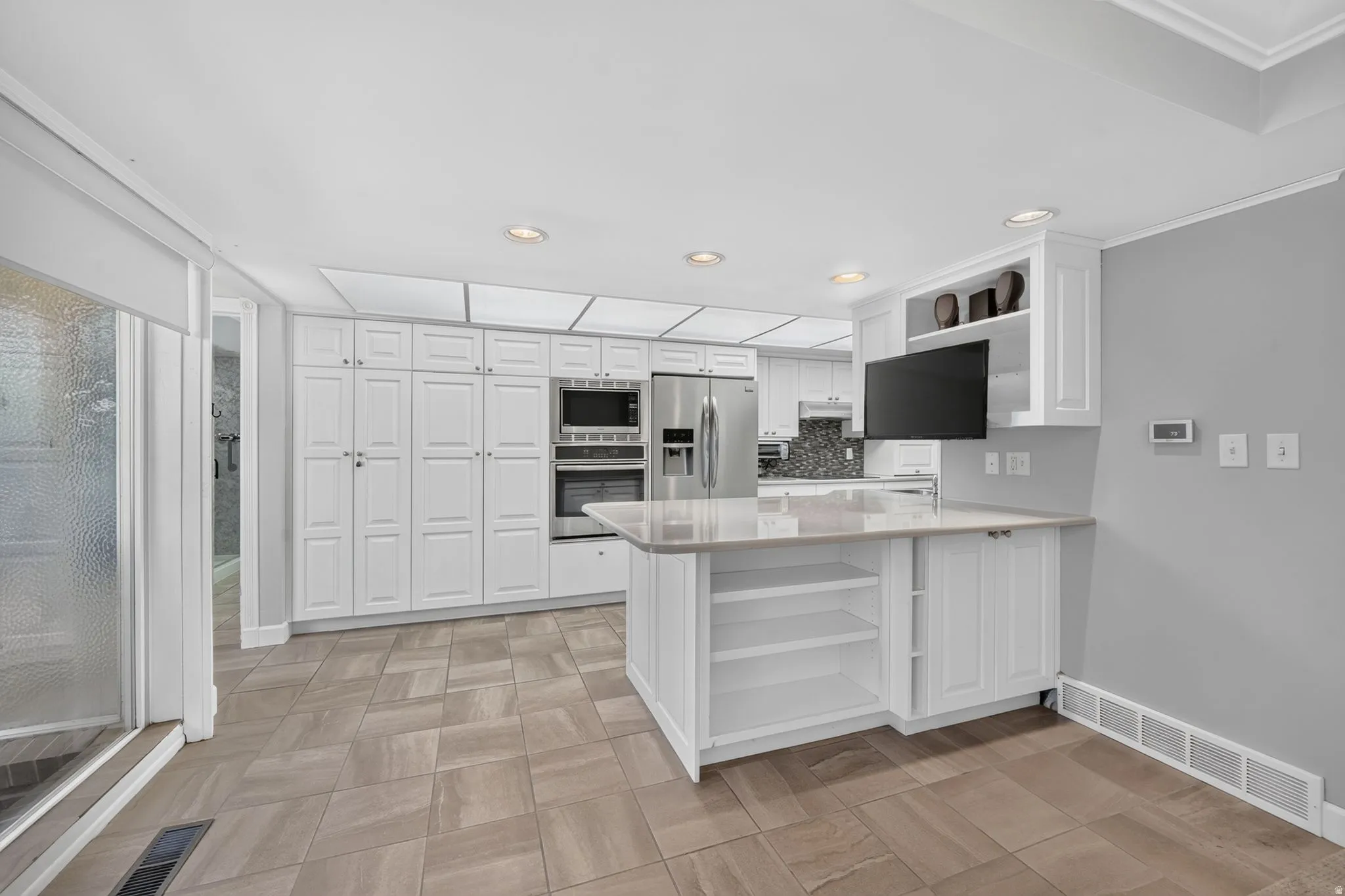 Contemporary kitchen featuring extensive white cabinetry, integrated stainless steel appliances, a center island with open shelving and countertop, and neutral-toned tile flooring