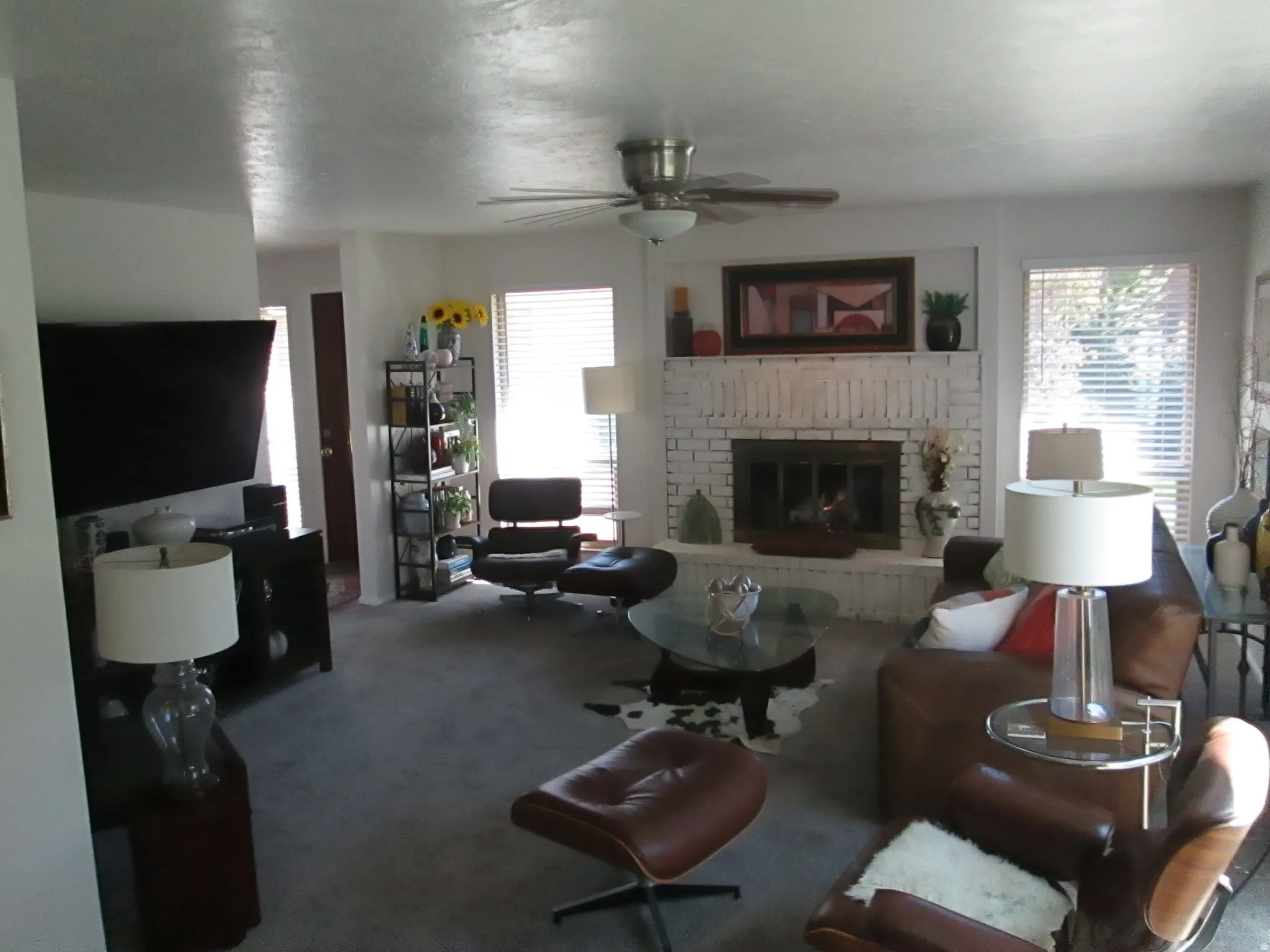 Living area featuring a painted brick fireplace, a ceiling fan with integrated lighting, and multiple windows with horizontal blinds
