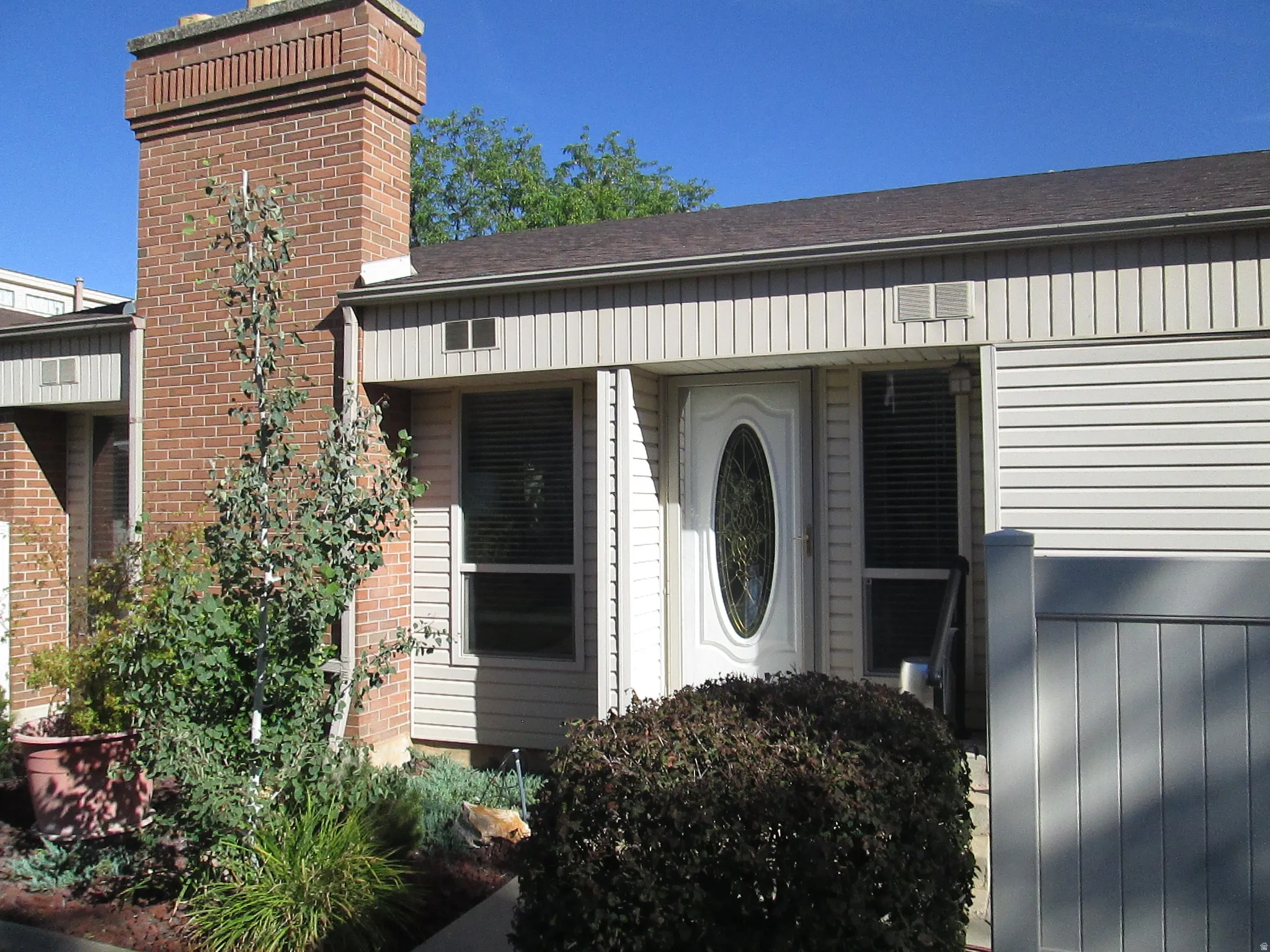 Brick chimney, neutral-toned siding, oval-paneled entry door, vertical privacy fencing, and landscaping with shrubs