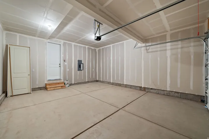 Concrete flooring with expansion joints, unfinished drywall walls, interior access door, overhead lighting, and an automatic garage door opener mechanism