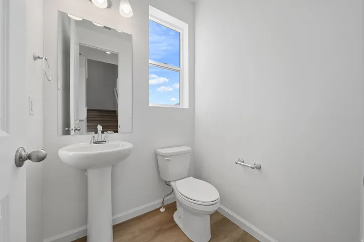 Pedestal sink with chrome faucet, wall-mounted mirror, natural light window, toilet, and wood-finish flooring