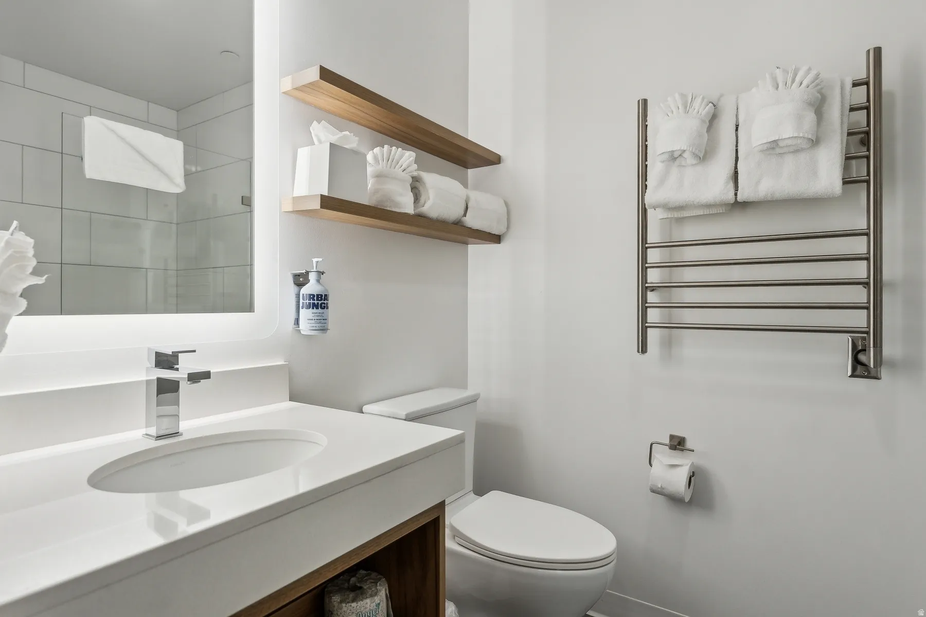 Vanity with integrated sink and chrome faucet, illuminated mirror, wood-finish floating shelves, and wall-mounted towel warmer