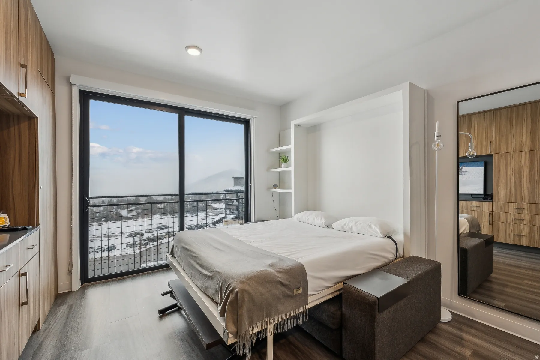 Wall-mounted bed with integrated shelving, wood-finish flooring, full-height sliding glass doors, built-in wood-finish cabinetry, and recessed lighting