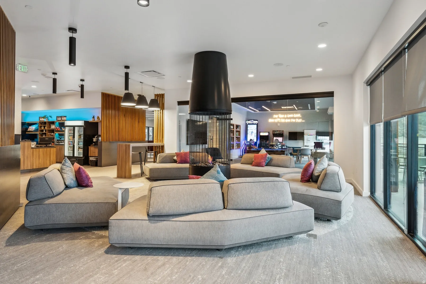 Spacious common area featuring textured flooring, a modern circular fireplace, a built-in refreshment bar, and expansive floor-to-ceiling windows with roller shades