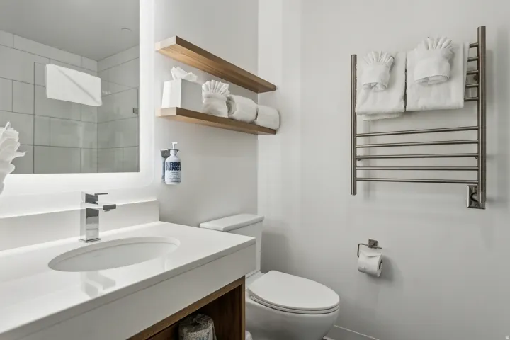 Vanity with integrated sink and chrome faucet, illuminated mirror, wood-finish floating shelves, and wall-mounted towel warmer