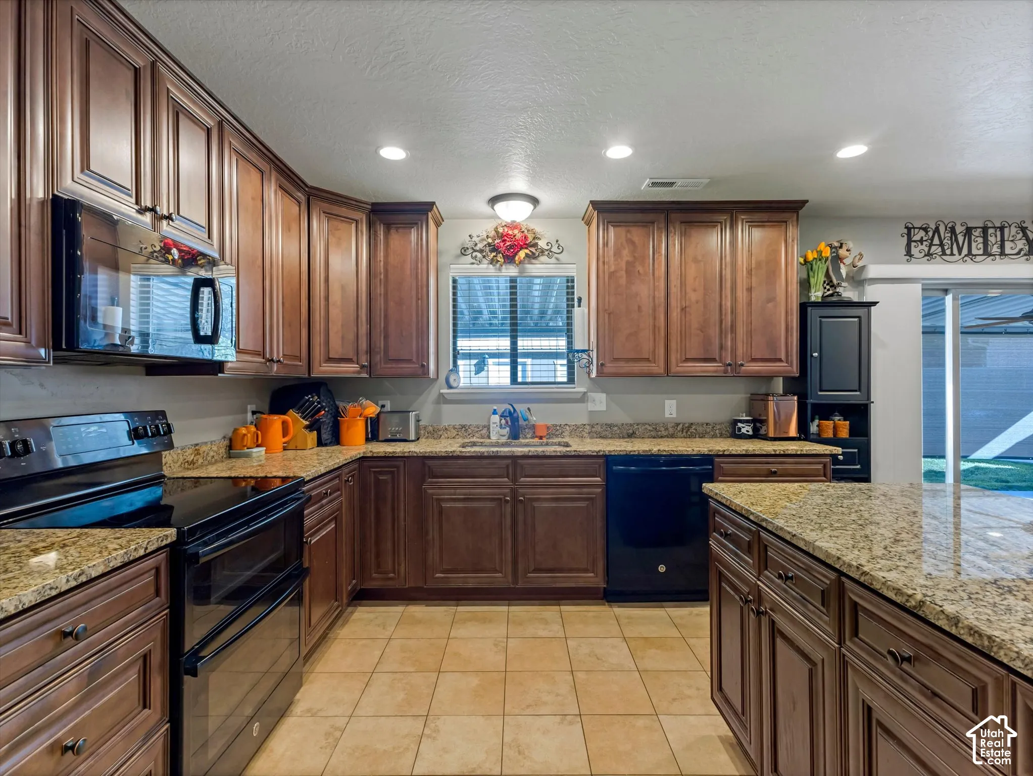 Brown wood-finish cabinetry, light-toned tiled flooring, dark built-in appliances, and stone-finish countertops