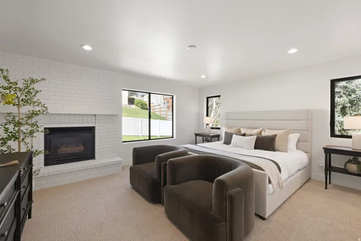 White brick fireplace with hearth, recessed lighting, expansive windows with dark frames, light-toned carpet, and a paneled upholstered headboard