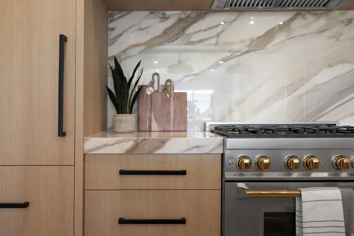 Wood-finish cabinetry with matte black hardware, stone countertops, stone slab backsplash, stainless steel range with gold-finish knobs, and integrated range hood