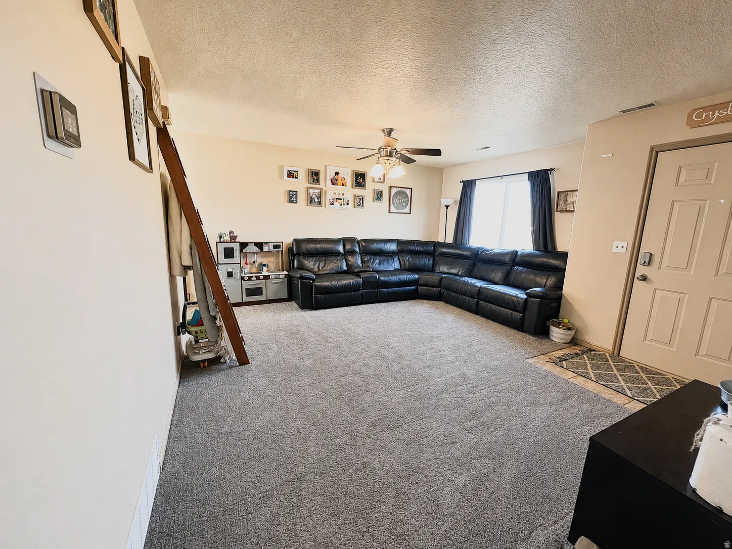 Carpeted living area featuring a ceiling fan with integrated lighting, a window with dark curtains, and a light-colored entry door