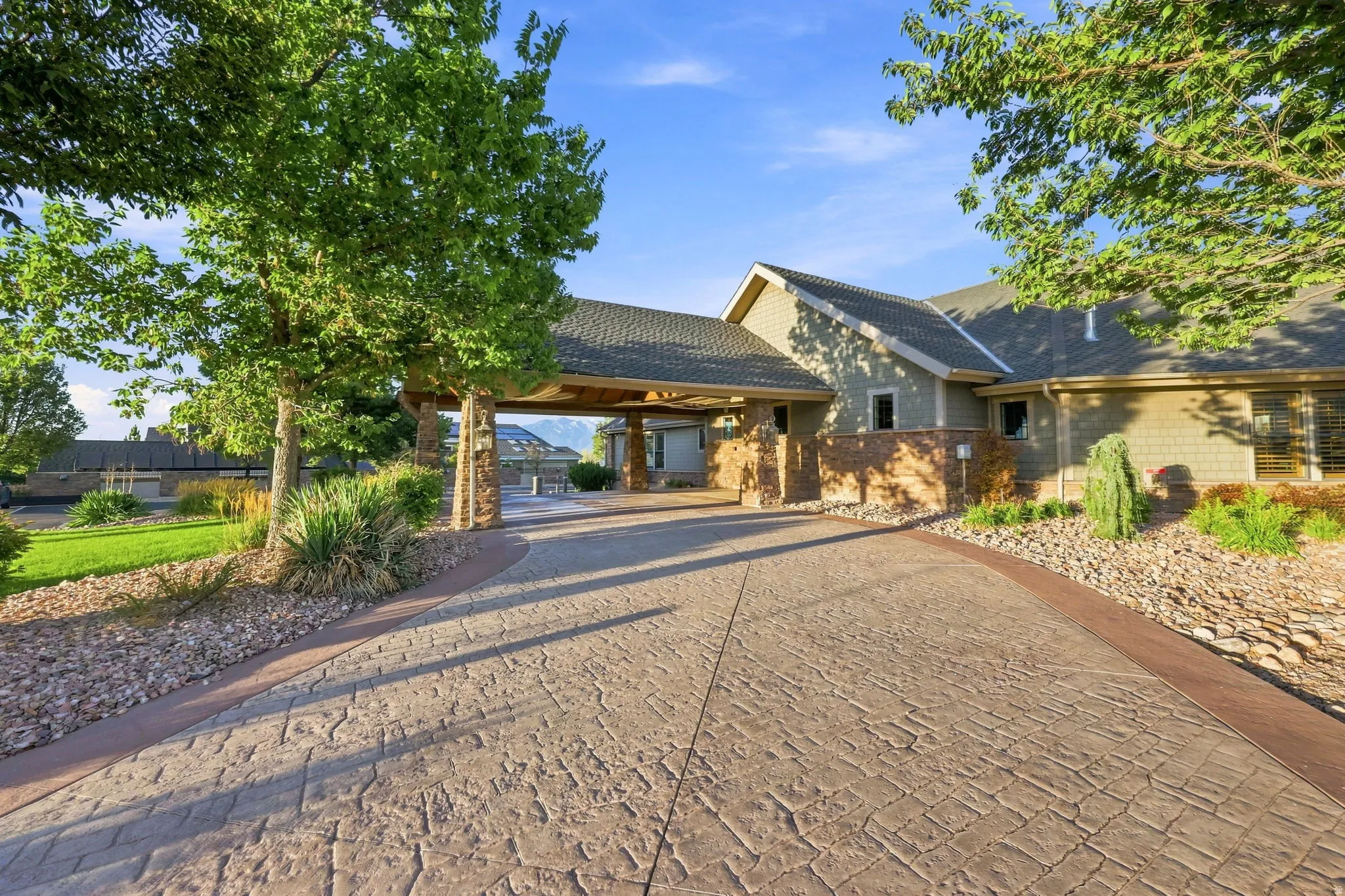 Textured concrete driveway, covered porte-cochere with stone columns, landscaped rock beds, and mature trees