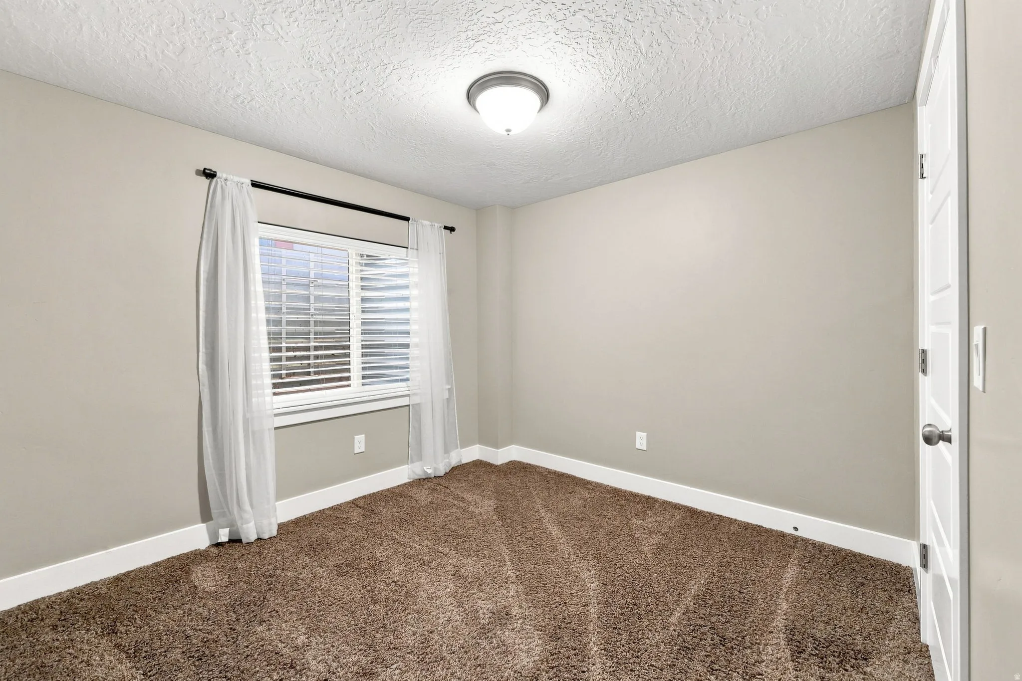 Neutral tone walls, textured ceiling, brown carpet, window with blinds, panel door