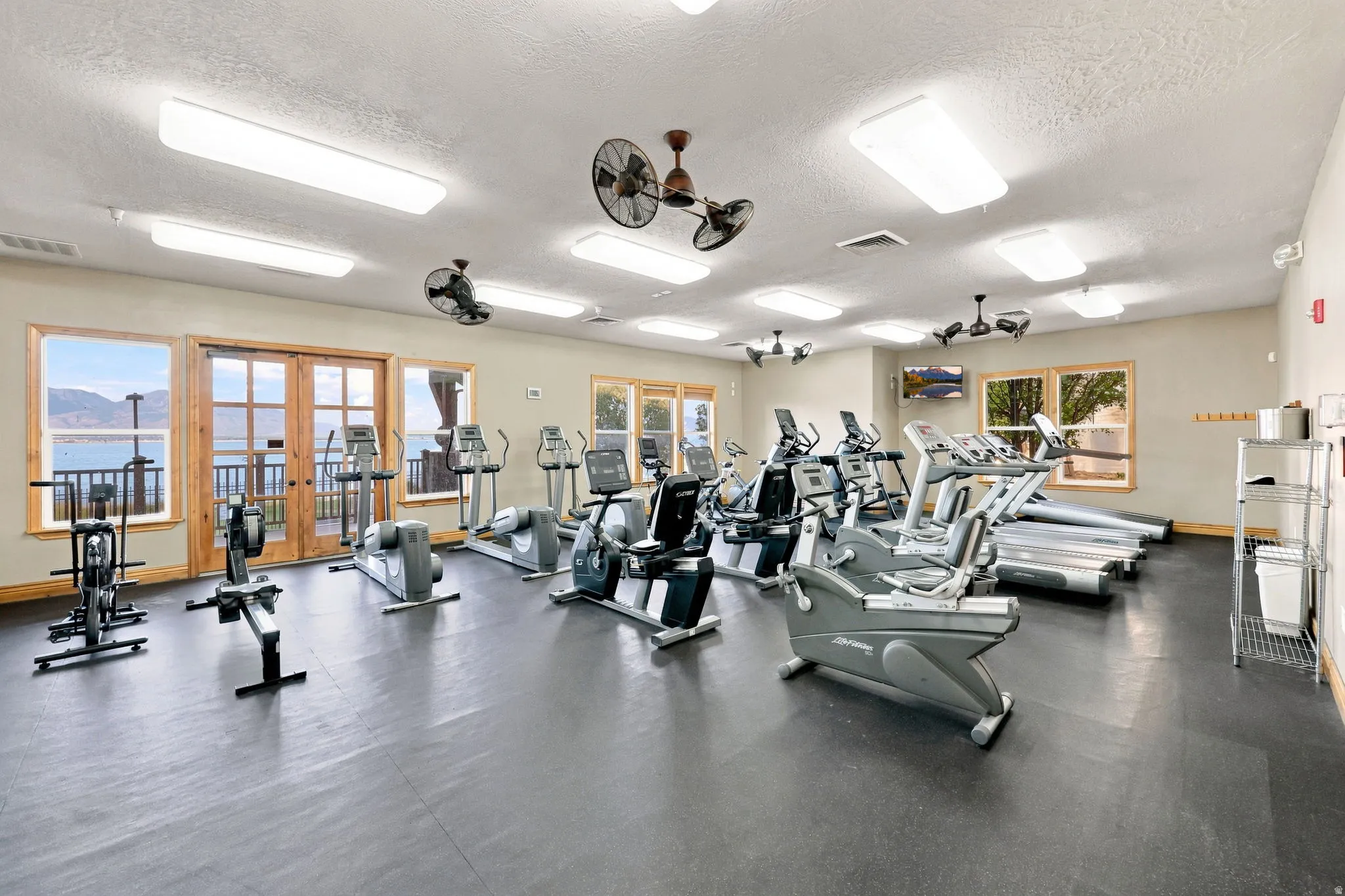 Fitness center with rubber flooring, multiple cardio machines, ceiling-mounted fans, and large windows with extensive water scenery