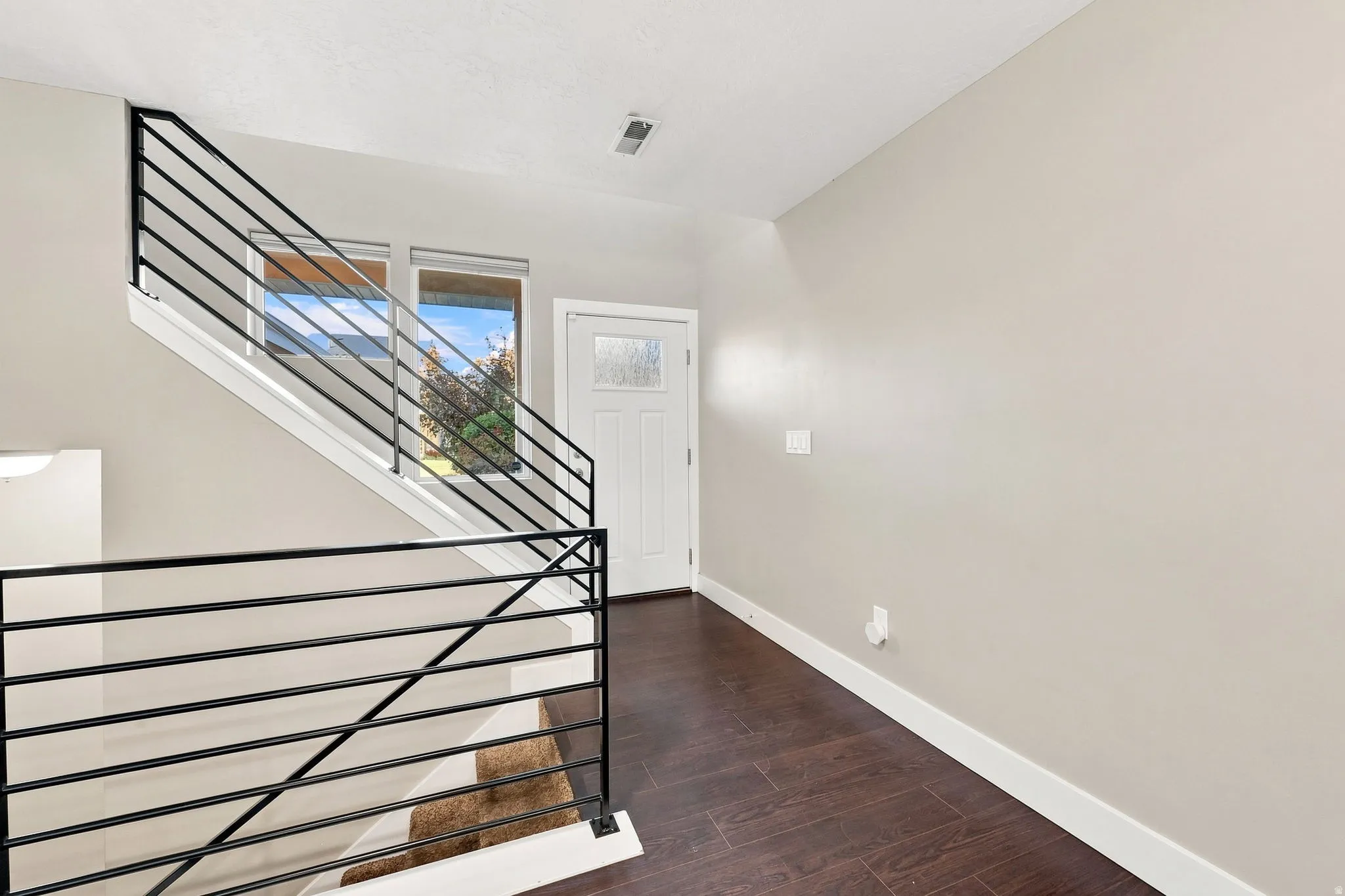 Dark wood-finish flooring, staircase with horizontal black metal railings, white panel door with decorative glass, and two upper windows
