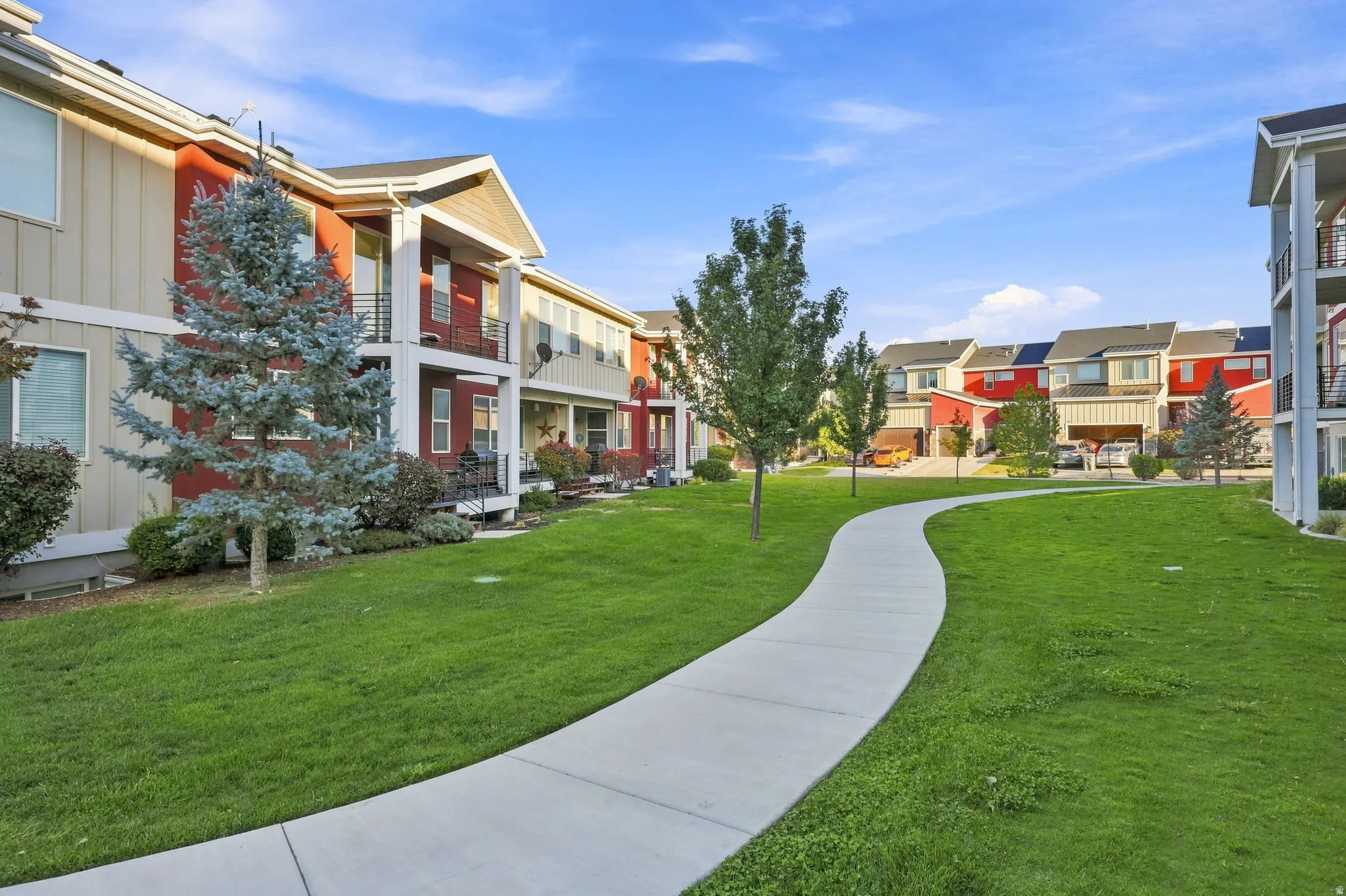 Manicured lawn with mature landscaping and curved concrete pathway, surrounded by multi-unit residential architecture featuring varied exterior finishes and pitched rooflines