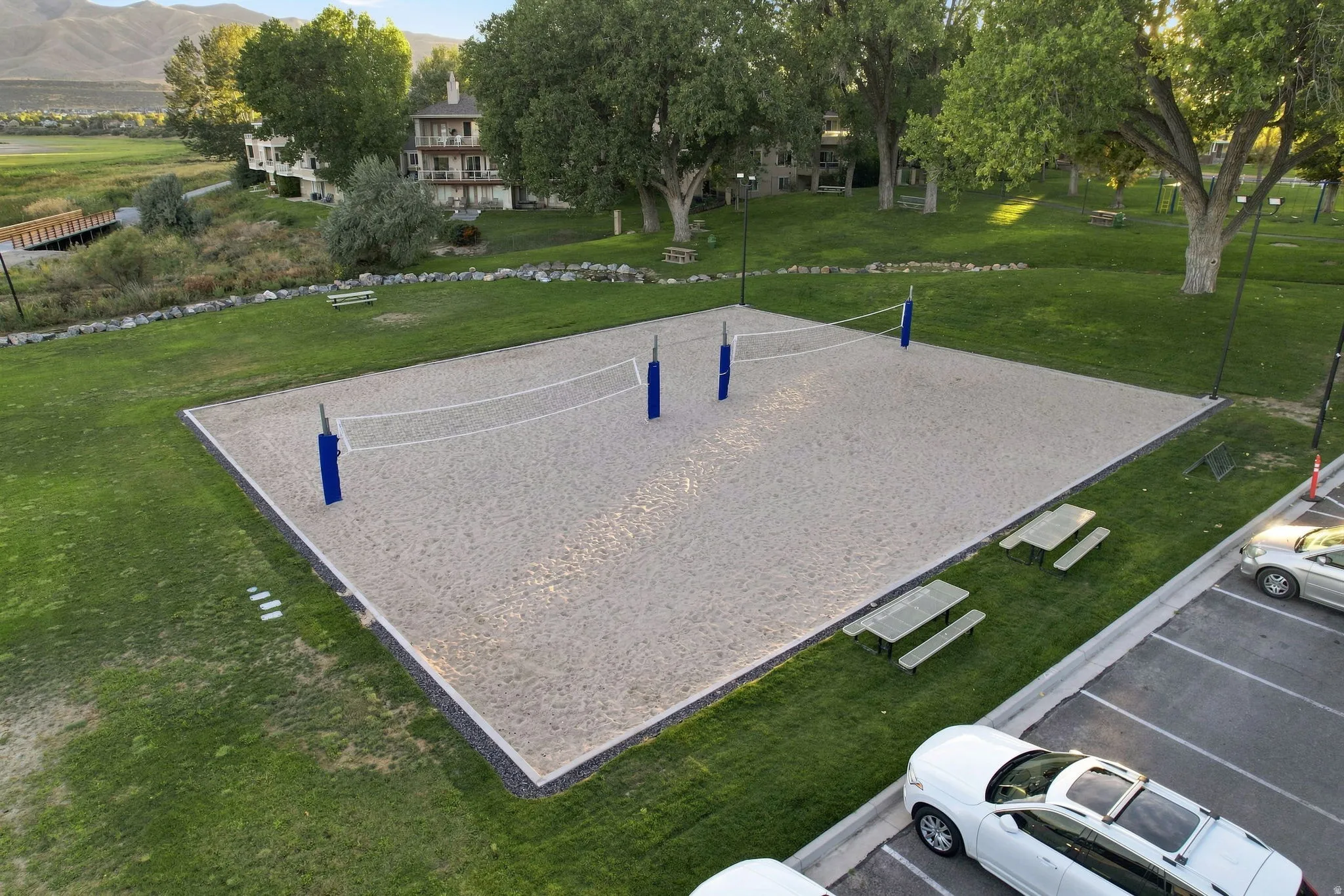 Sand volleyball court with blue net posts, surrounded by green lawn and mature trees