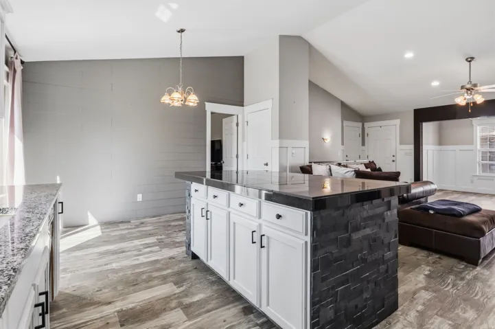 Open-concept interior featuring wood-finish flooring, vaulted ceilings, a kitchen island with stone veneer, and white cabinetry with dark hardware