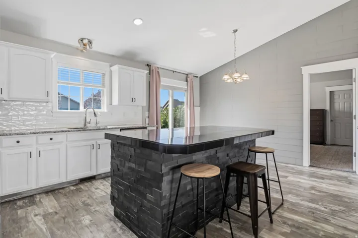 Vaulted ceiling, wood-finish flooring, white cabinetry, tile backsplash, and a stone-clad kitchen island