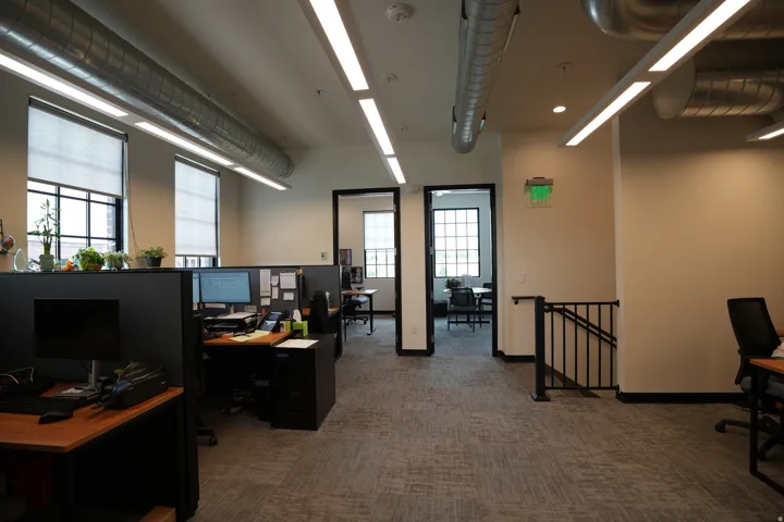 Open-concept office space featuring exposed ductwork, suspended linear lighting, large windows with roller shades, and carpet tile flooring